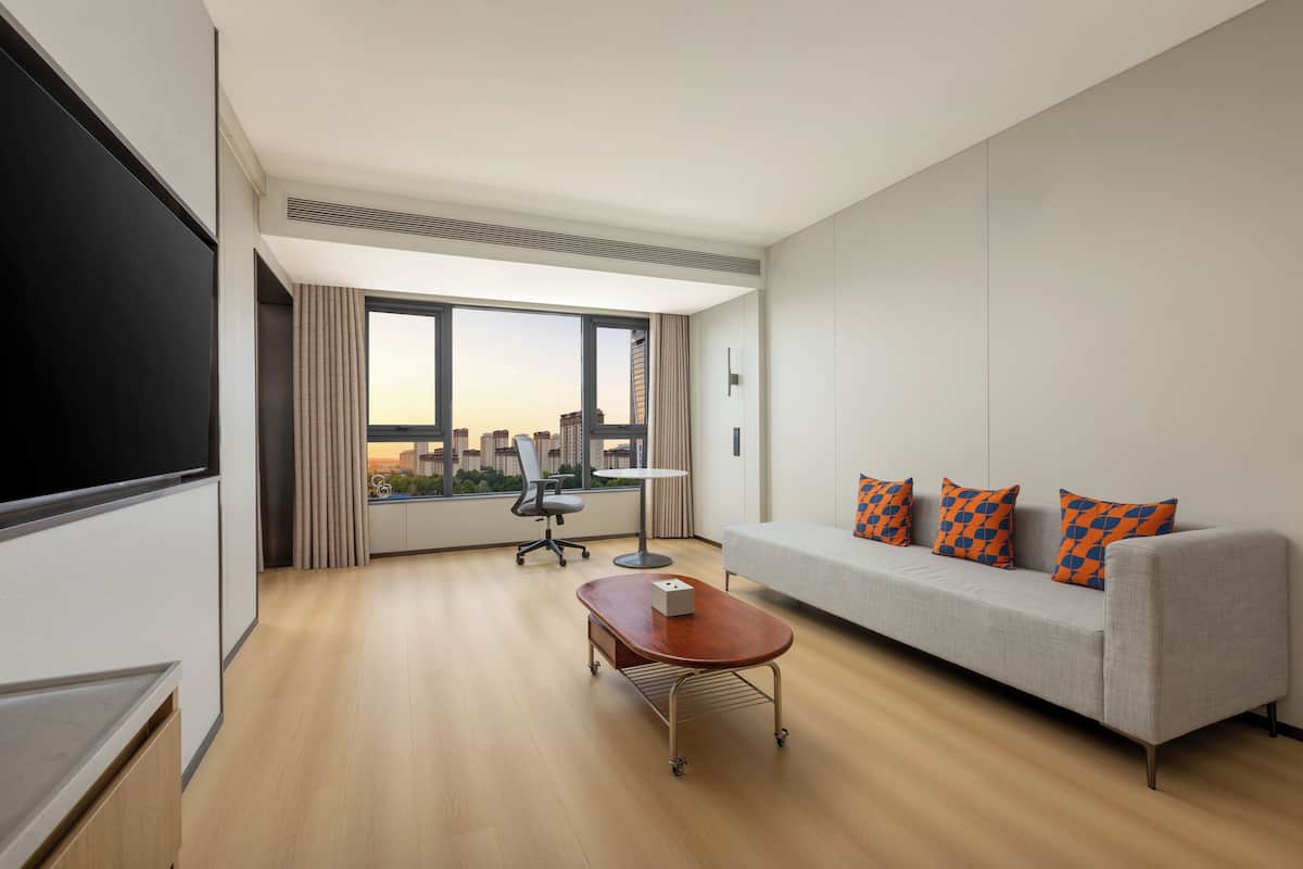 A modern hotel room with a large window, a sofa, a desk, and a flat-screen TV.