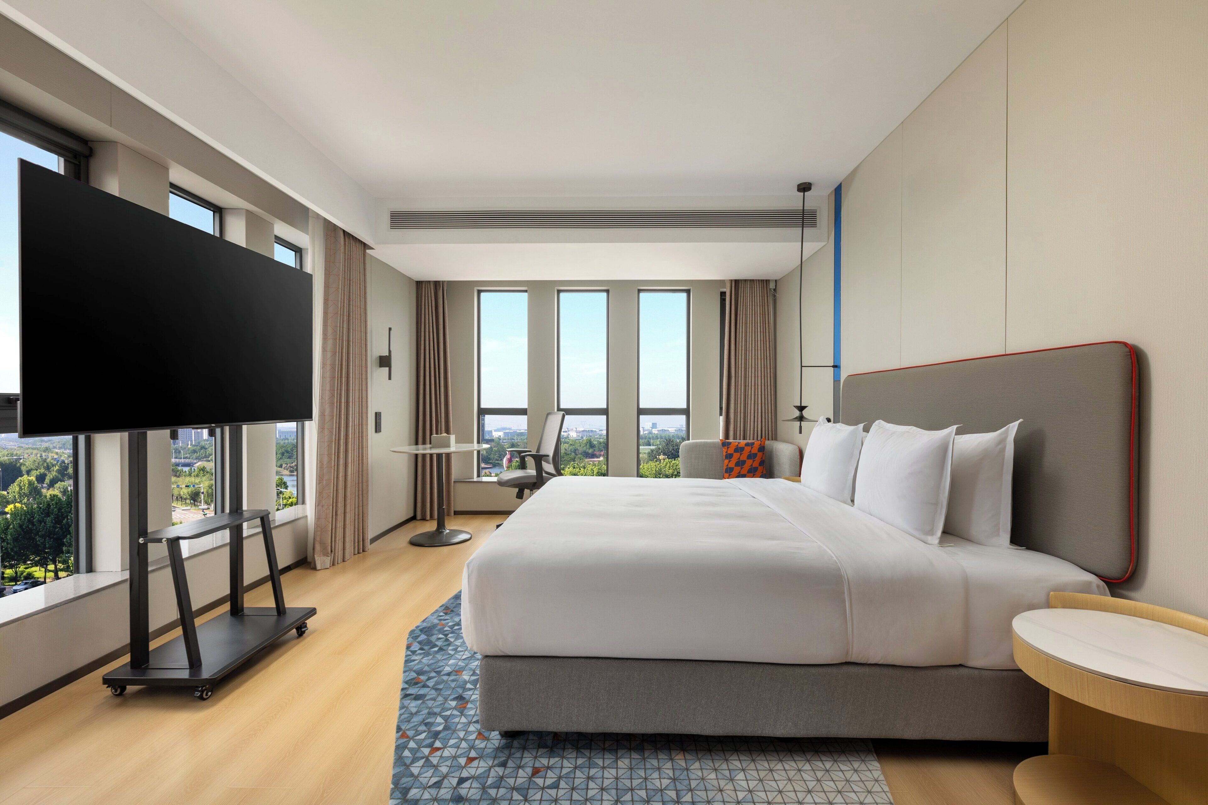 A modern hotel room with a large bed, a desk, a flat-screen TV, and a view of the city.