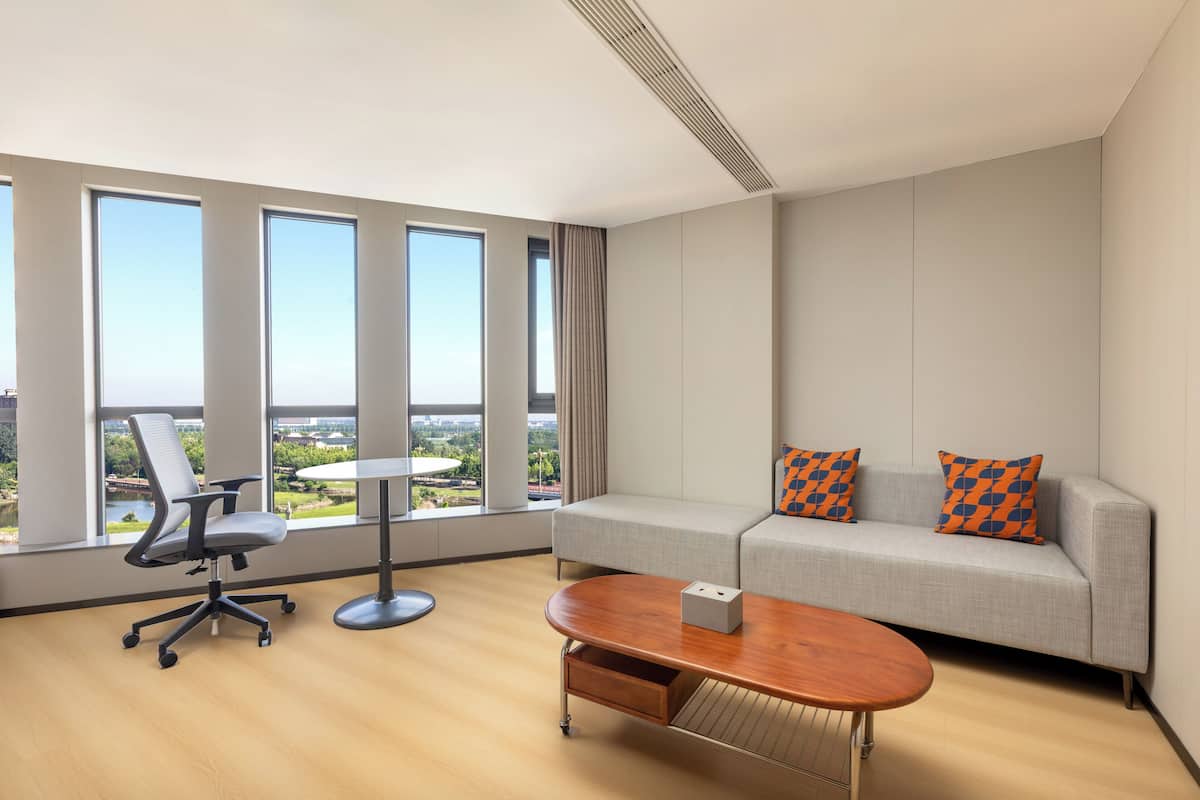 A modern hotel room with a large window, a grey sofa with orange patterned pillows, a wooden coffee table, and a grey office chair.
