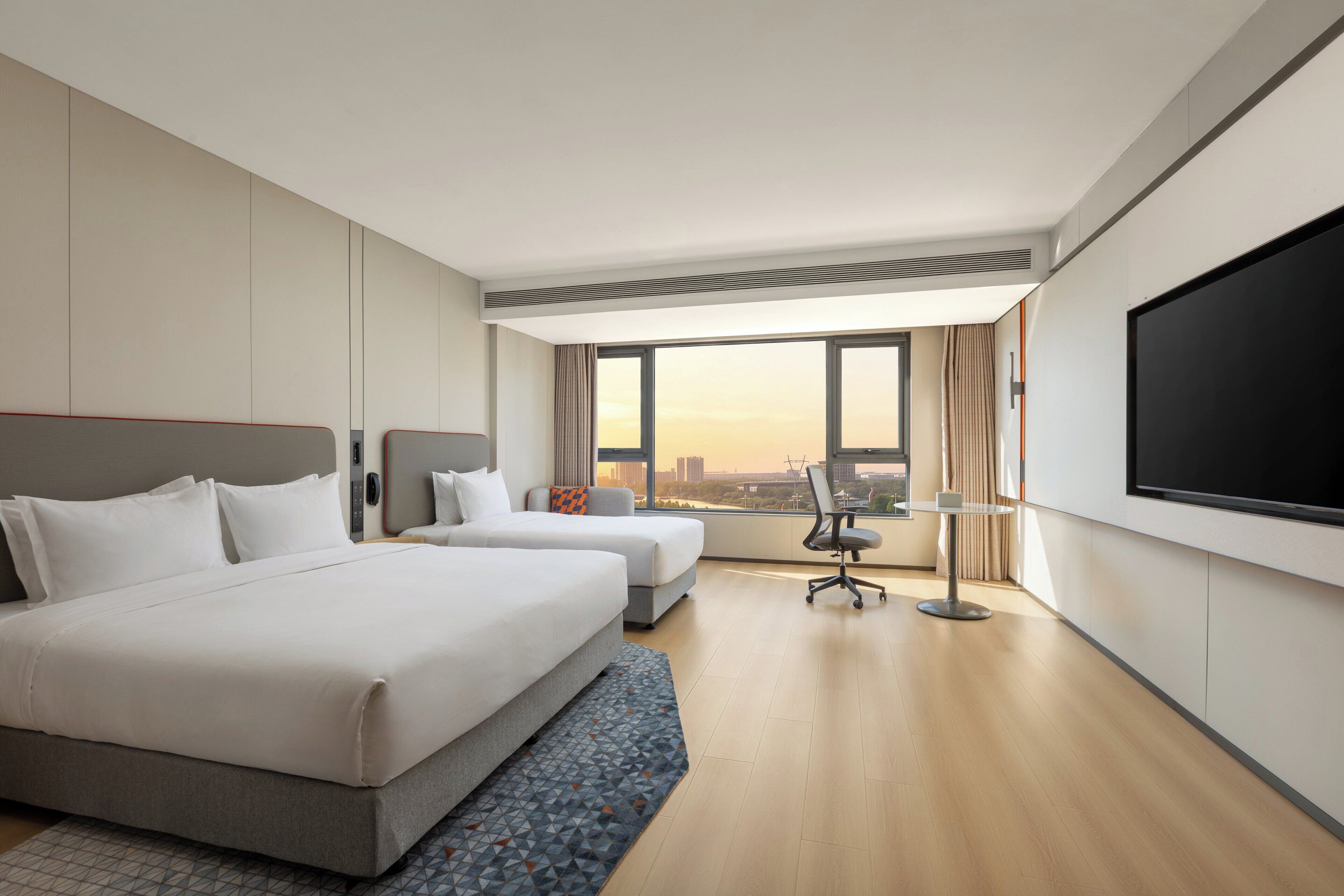 A hotel room with two beds, a desk, a chair, a large TV, and a view of the cityscape.