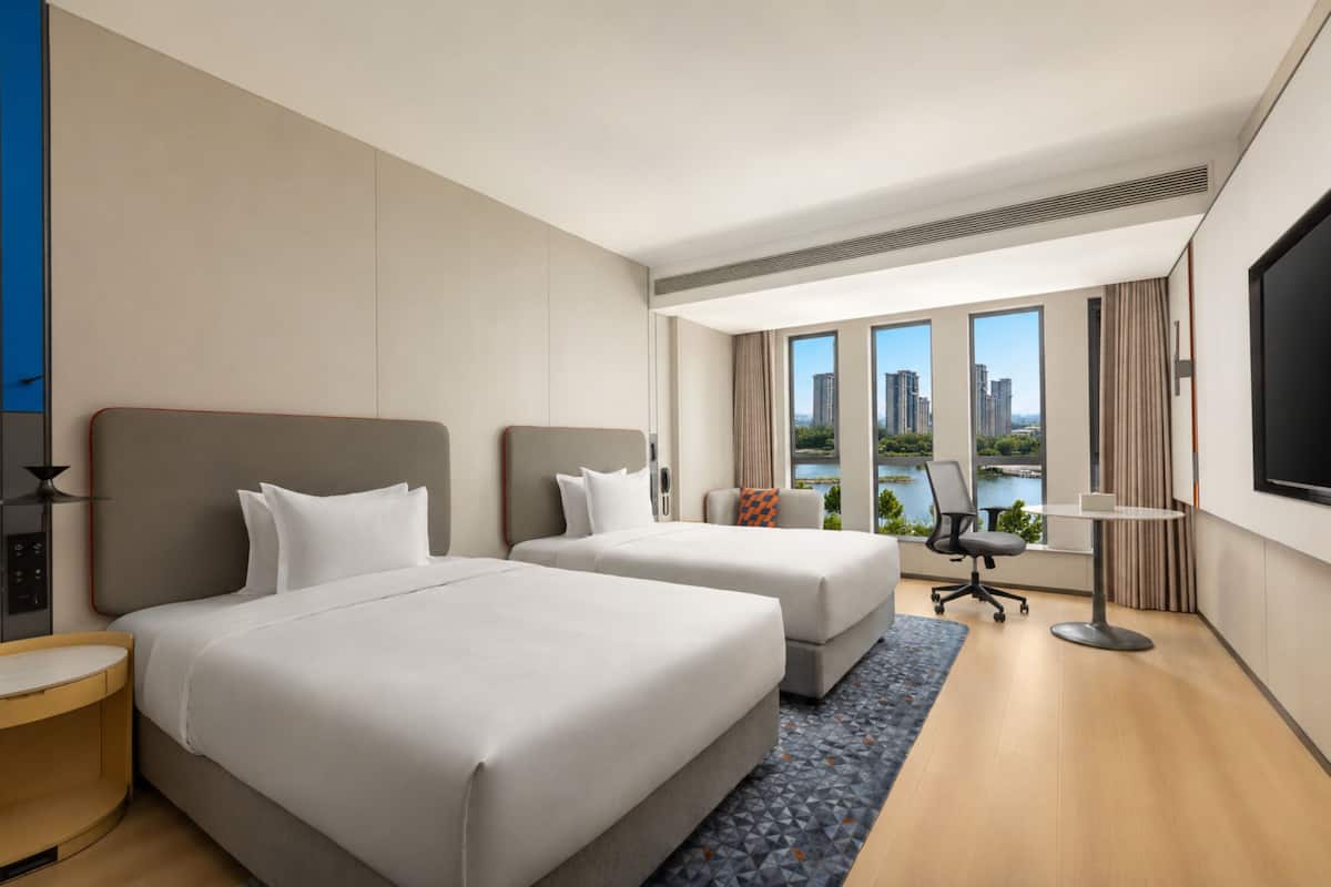 A hotel room with two beds, a desk, a chair, a TV, and a large window with a city view.