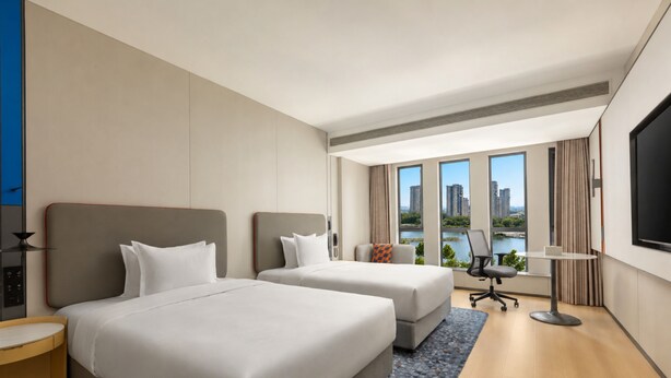 Ein Hotelzimmer mit zwei Betten, einem Schreibtisch, einem Stuhl, einem Fernseher und einem großen Fenster mit Stadtblick.