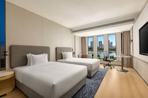 A hotel room with two beds, a desk, a chair, a TV, and a large window with a city view.