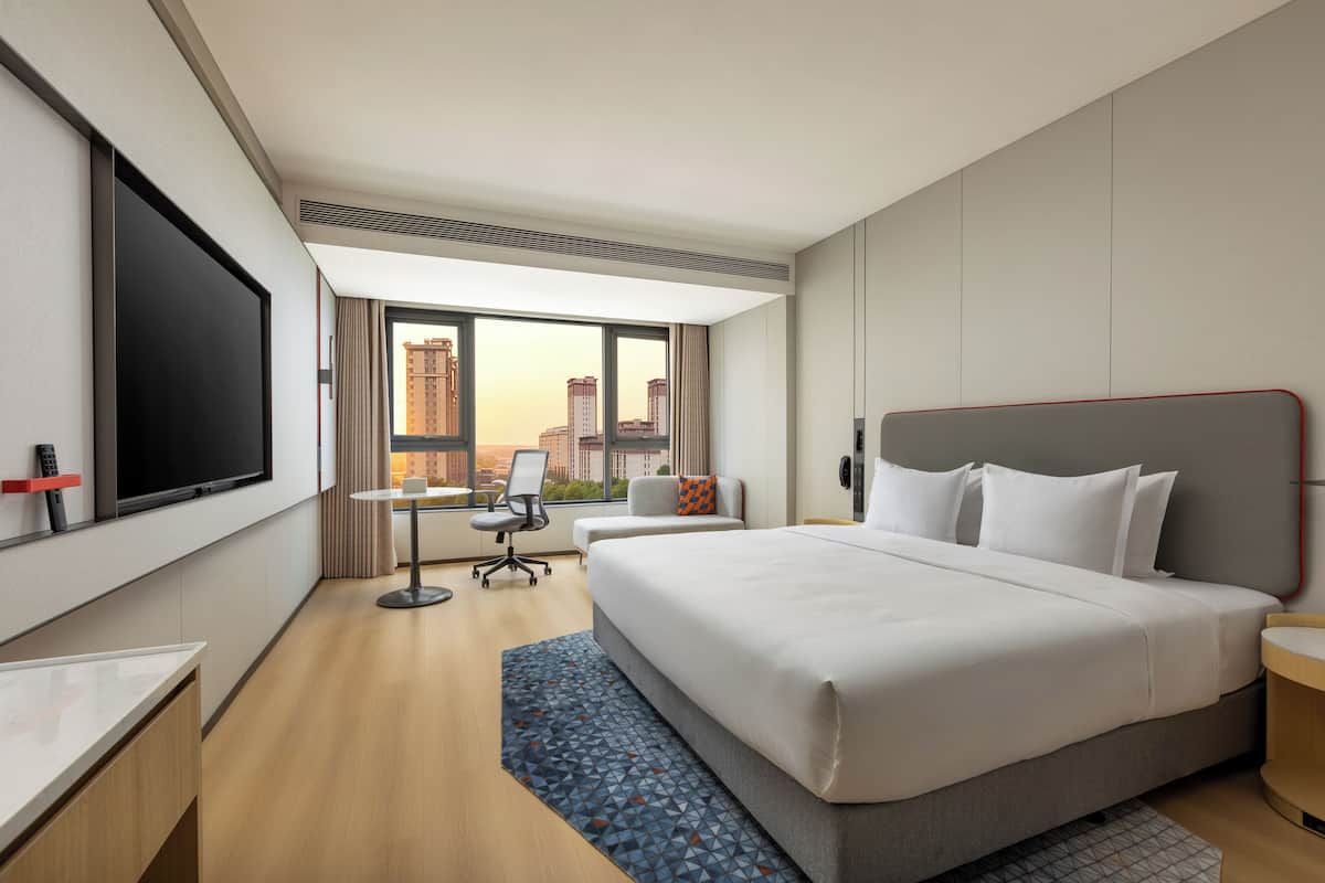A hotel room with a large bed, a desk with a chair, a TV, and a view of tall buildings.