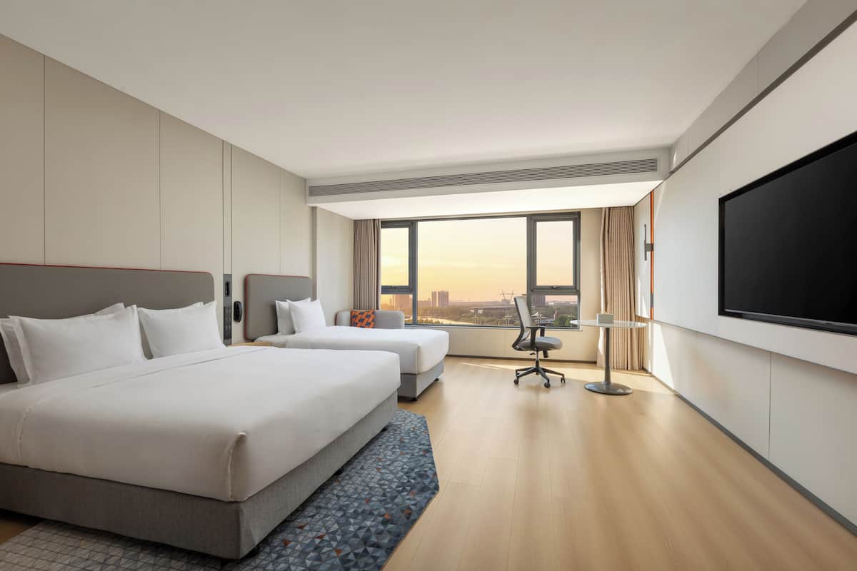 A hotel room with two beds, a TV, a desk, and a large window with a city view.