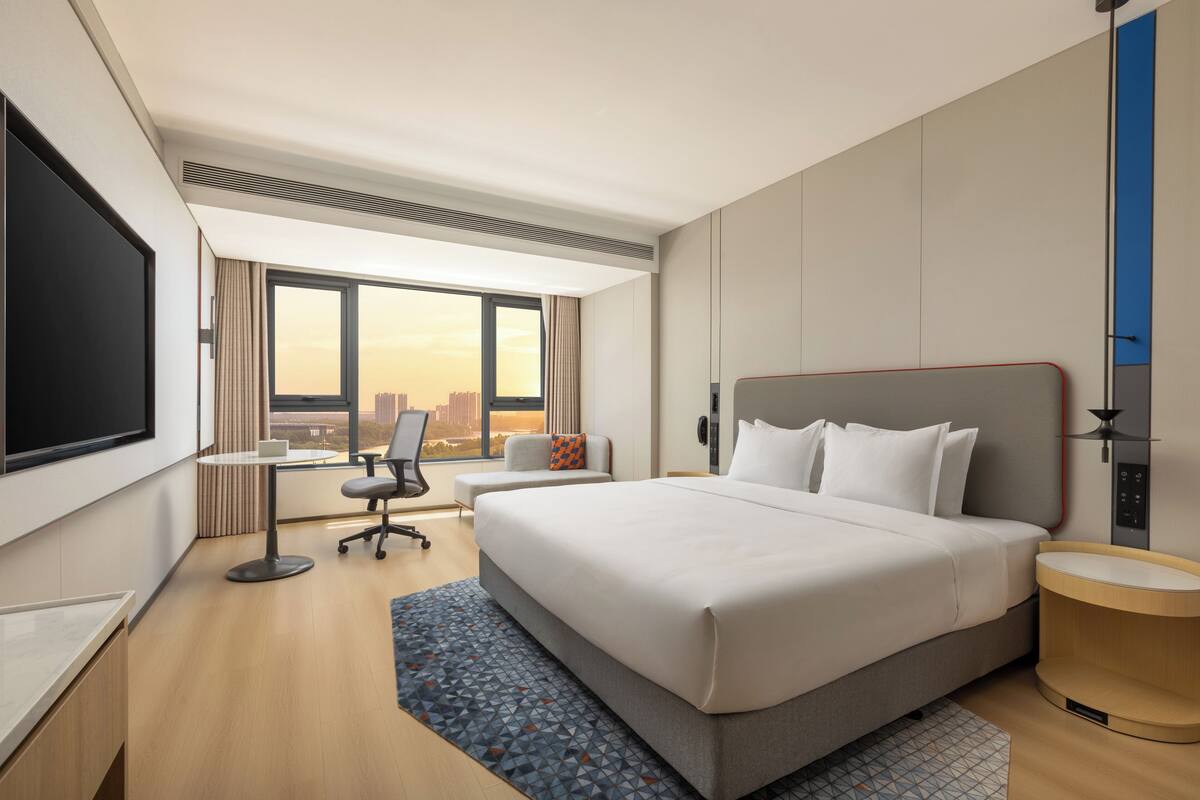 A hotel room with a large bed, a desk, a chair, a TV, and a window with a city view.