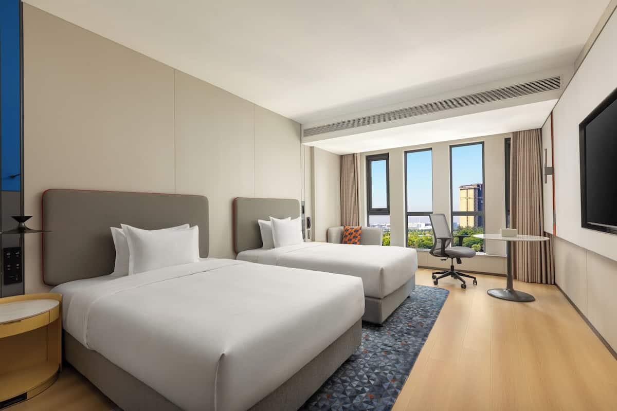 A hotel room with two beds, a desk, a chair, a TV, and large windows.