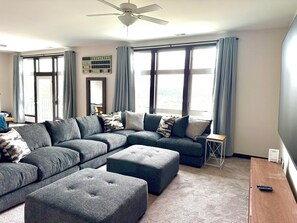 A living room with a sectional sofa, ottomans, a small side table, and a ceiling fan. - The Dugout - Baseball Oasis Duplex Condo (Dyersville)