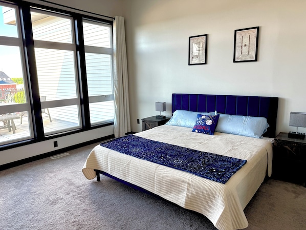A bedroom with a large window, a bed with a blue headboard, two framed pictures on the wall, and a nightstand with a lamp. - The Dugout - Baseball Oasis Duplex Condo (Dyersville)