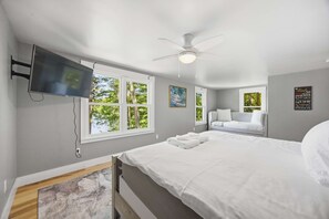 Een slaapkamer met een bed, een plafondventilator, een aan de muur bevestigde televisie en een raam met uitzicht op bomen.
