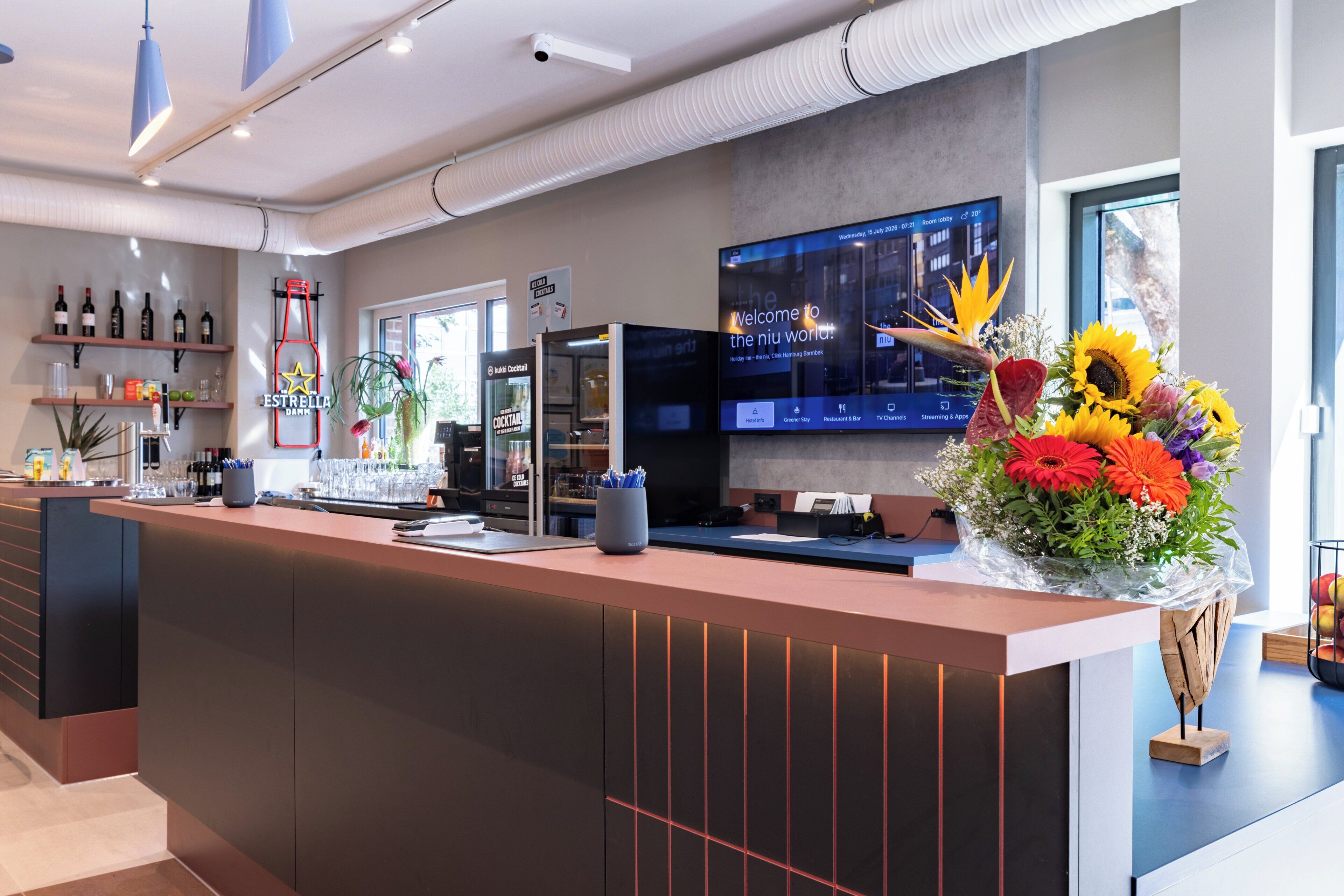 Un mostrador de recepción de hotel con una pantalla digital mostrando información de las habitaciones, diversas bebidas embotelladas en estantes y un arreglo floral.