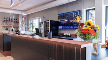 Un mostrador de recepción de hotel con una pantalla digital mostrando información de las habitaciones, diversas bebidas embotelladas en estantes y un arreglo floral.