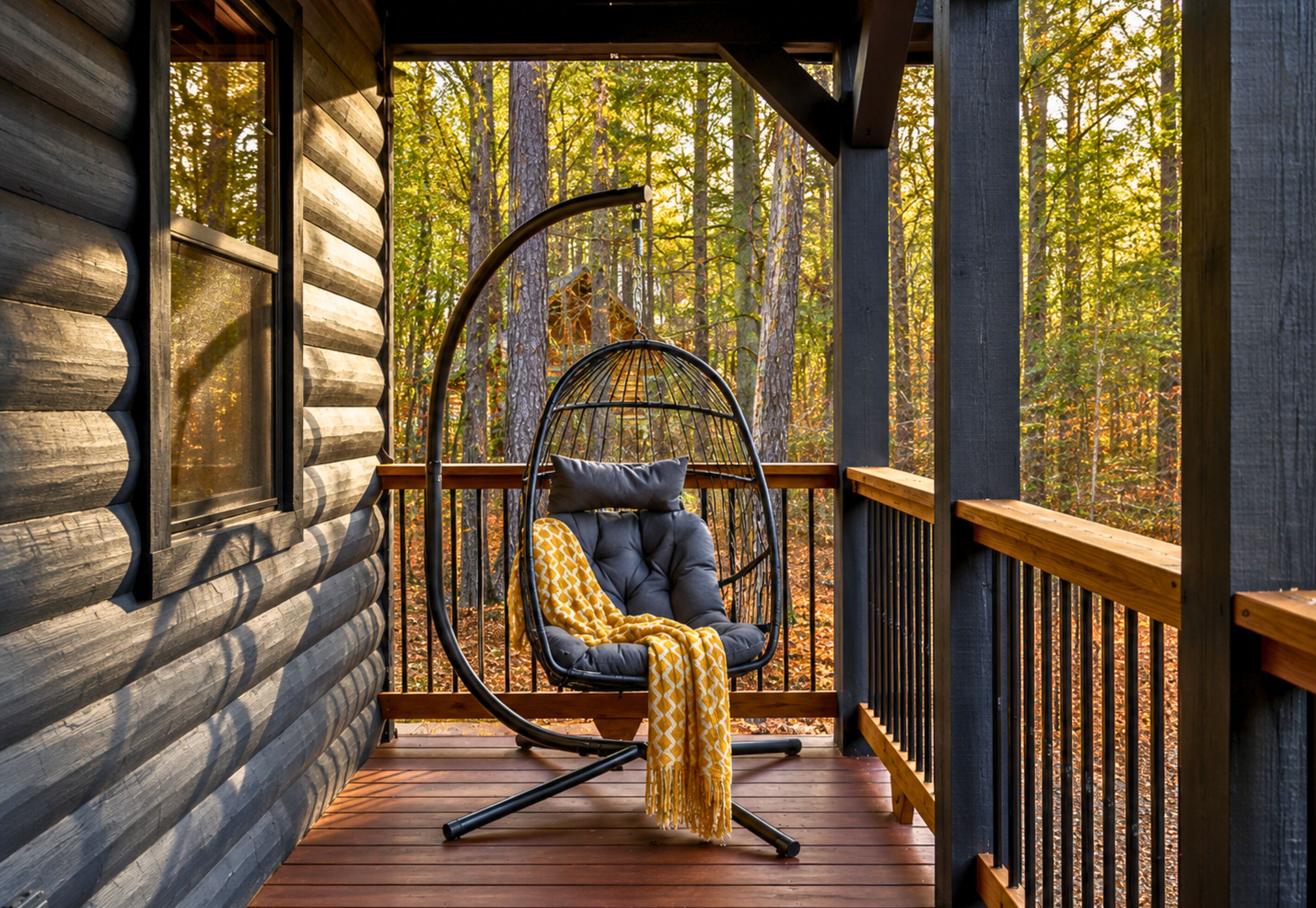 Une cabane en rondins avec une terrasse en bois, un fauteuil suspendu et une vue sur une forêt.