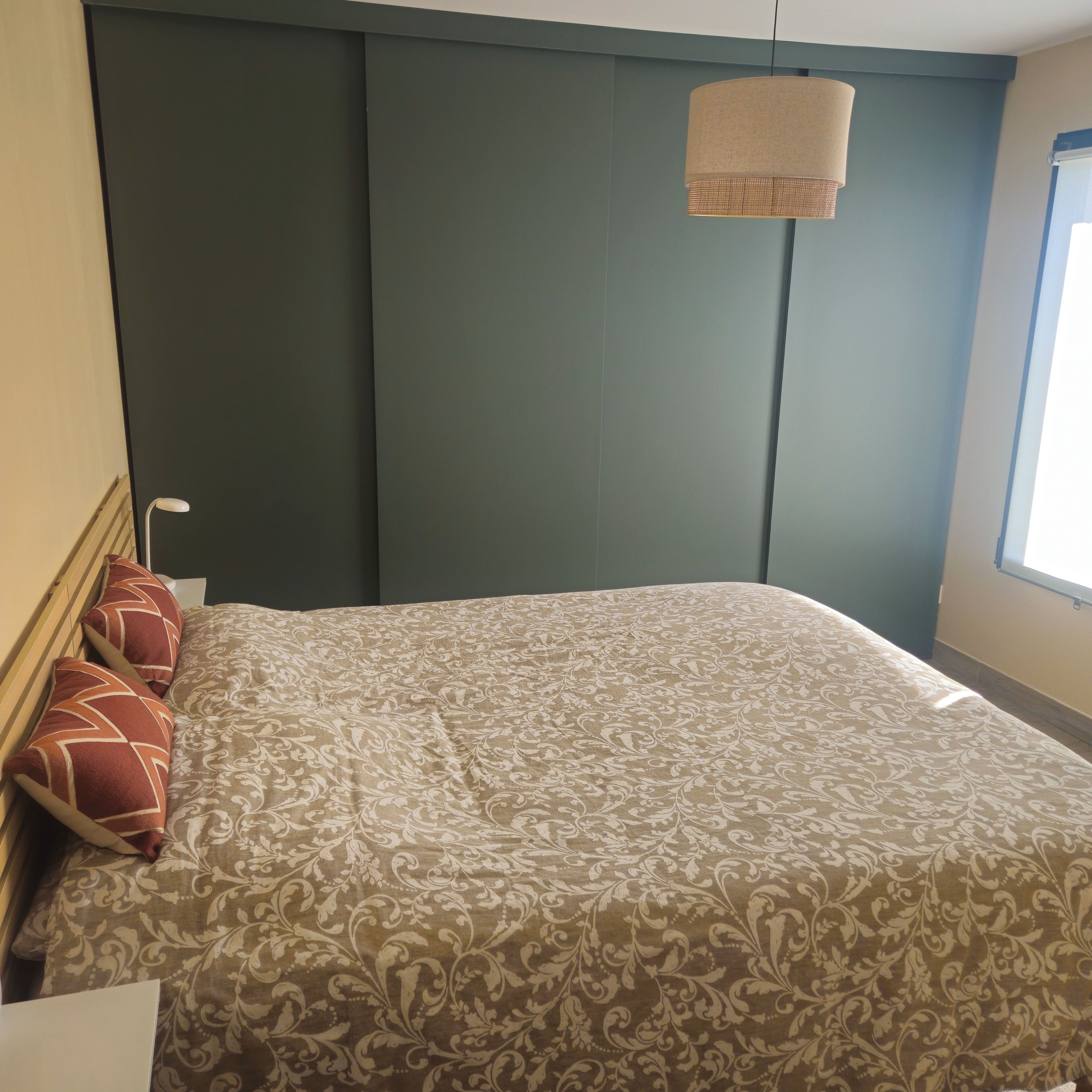 A bed with a patterned bedspread, a hanging light fixture, and a window with blinds.