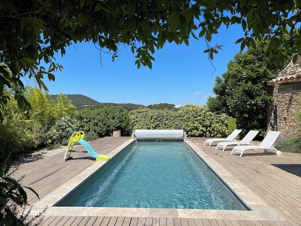 Une piscine rectangulaire avec un toboggan, entourée d’une terrasse en bois et agrémentée de chaises longues blanches.