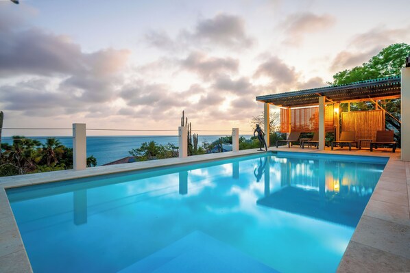 A swimming pool with a view of the sea, a covered seating area, and a staircase leading to a wooden deck. - Casa Doce – Charming Ocean View Villa with Private Pool (Kralendijk)
