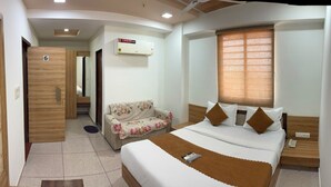 A bedroom with a bed, bedside table, nightstand, and a window with blinds. - Hotel Yuvraj (Gandhinagar)