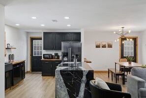 A modern kitchen with dark cabinets, a marble island, and a dining area with a table and chairs. - Huckleberry Arch - Cozy RRG Home near Natural Arch (Pine Ridge)