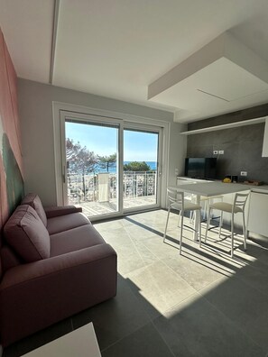 Un salon moderne avec un canapé, un coin repas avec des chaises, une cuisine avec un téléviseur et un balcon offrant une vue sur la mer.