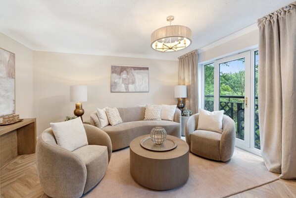 A living room with a sectional sofa, two armchairs, a round coffee table, a chandelier, and a large window with curtains.
