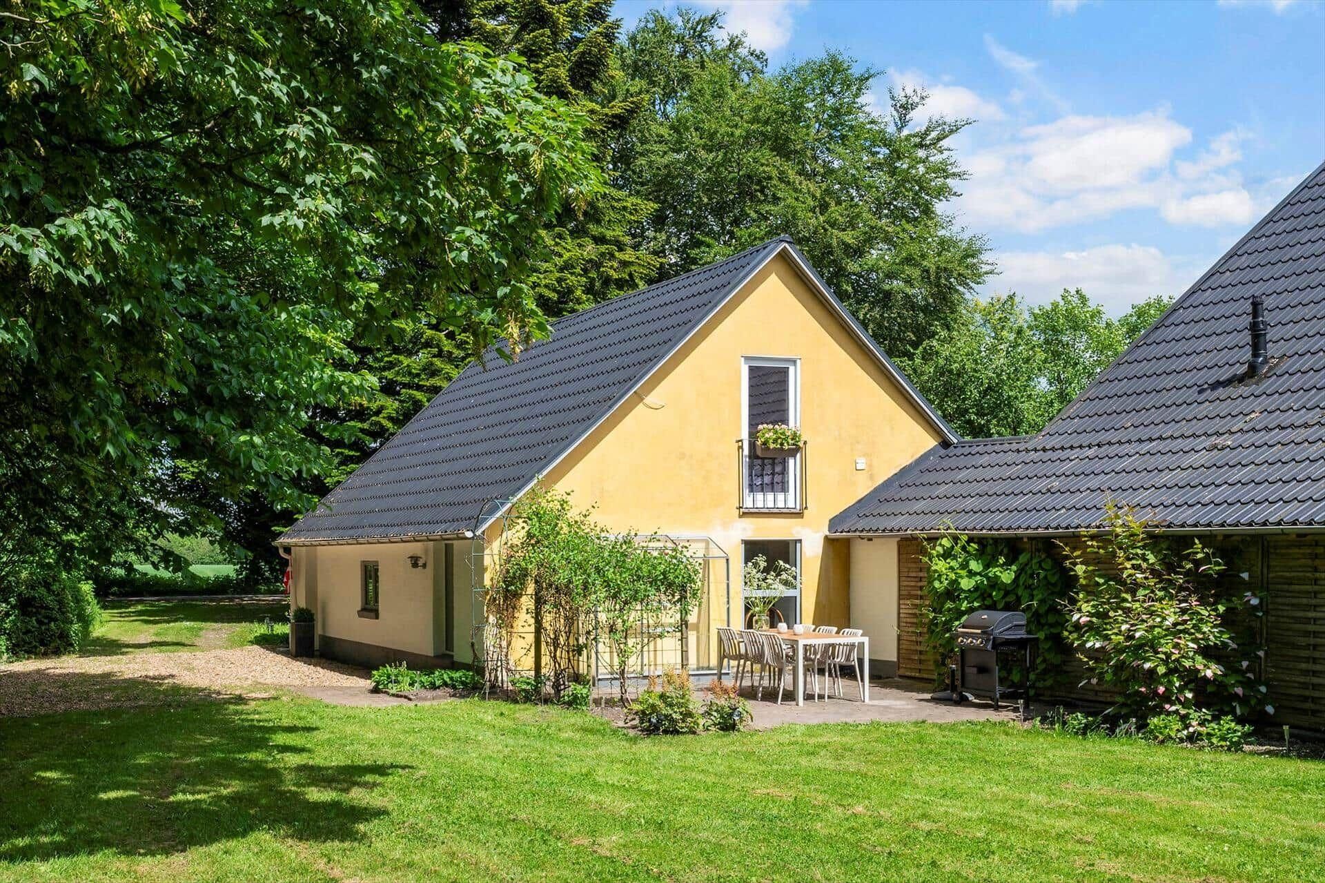 A yellow house with a sloped roof, a small balcony with plants, and a patio with a table and chairs.