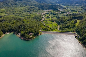 Eine Vogelperspektive auf einen See, der von bewaldeten Hügeln, einem Stausee und einer kleinen Siedlung mit sichtbaren Straßen und Gebäuden umgeben ist.