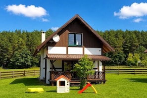Una casa de dos pisos con un balcón, una casita de juegos y un tobogán en un césped.