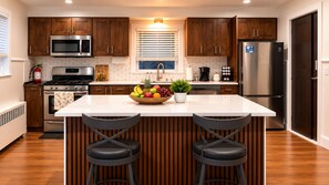 A modern kitchen with wooden cabinets, stainless steel appliances, a central island with bar stools, and a fruit bowl. -     Stylish 3BR Retreat Minutes from NYC & World Cup Events (City of Orange)