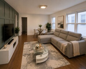 Ein Wohnzimmer mit Ecksofa, Glastisch, Flachbildfernseher, Essbereich mit Tisch und Stühlen sowie großem Fenster mit Aussicht.