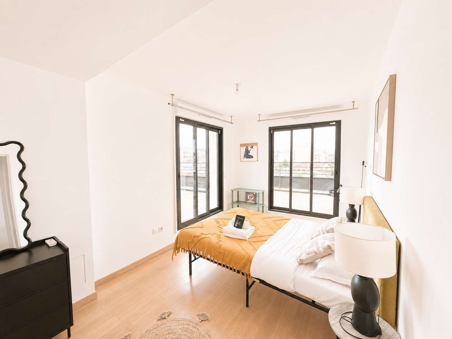 Une chambre à coucher comprenant un lit, une commode, une lampe et deux fenêtres donnant sur la ville.