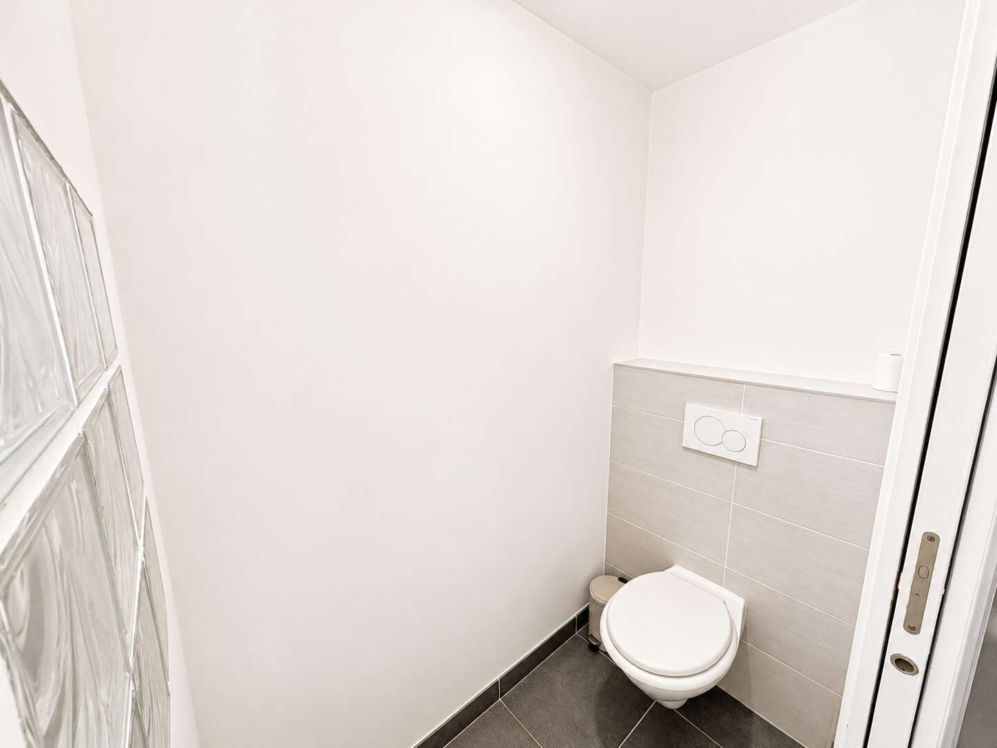 Ein kleines, sauberes Badezimmer mit Toilette, einem an der Wand montierten Spülmechanismus und einem gefliesten Boden.