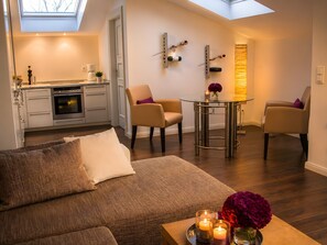Ein modernes Wohnzimmer mit einer Couch, zwei Sesseln, einem Couchtisch mit Kerzen und einer Küche im Hintergrund.
