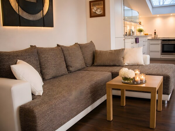 Ein modernes Wohnzimmer mit einer Eckcouch, einem Couchtisch mit Blumen und einer Küche im Hintergrund.