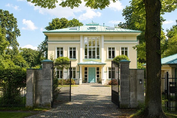 A large, two-story building with a green roof, a central entrance with a green door, and a paved driveway leading to a gated entrance.