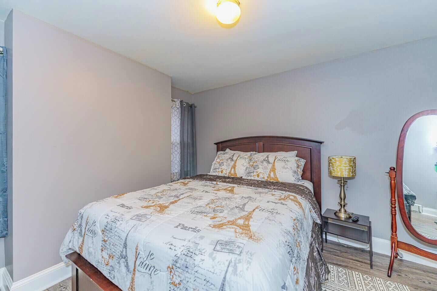 Une chambre à coucher avec un lit, une table de chevet, une lampe, un miroir et une fenêtre avec des rideaux.