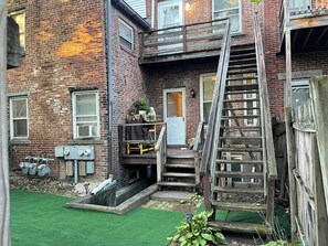 Un bâtiment en briques avec un escalier en bois menant à une porte, un petit espace extérieur avec une pelouse verdoyante et des boîtiers techniques.