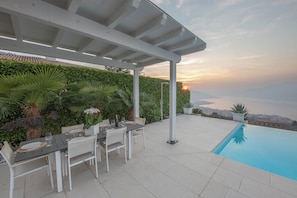 Um pátio externo com piscina, móveis brancos e vista para o pôr do sol sobre a água.
