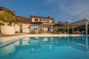 Piscina com espreguiçadeiras e guarda-sóis, uma grande planta em vaso e uma casa de dois andares com varanda.