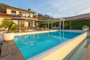 Piscina com espreguiçadeiras e guarda-sóis, um alpendre coberto e uma casa com varanda.