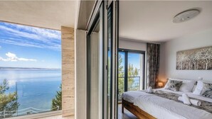 Une chambre avec un grand lit, un tableau au mur et une vue sur la mer, visible à travers une porte coulissante.