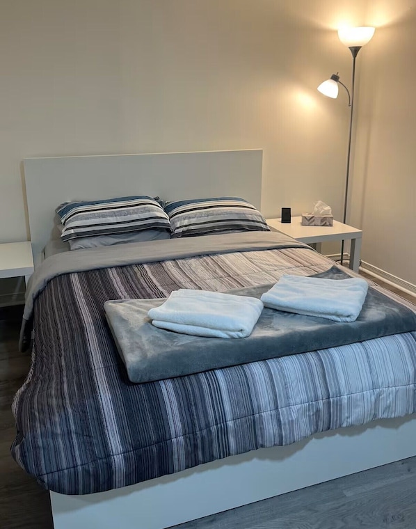 A neatly made bed with striped bedding, a bedside table with a lamp, and a small bedside table with a tissue box and a phone.