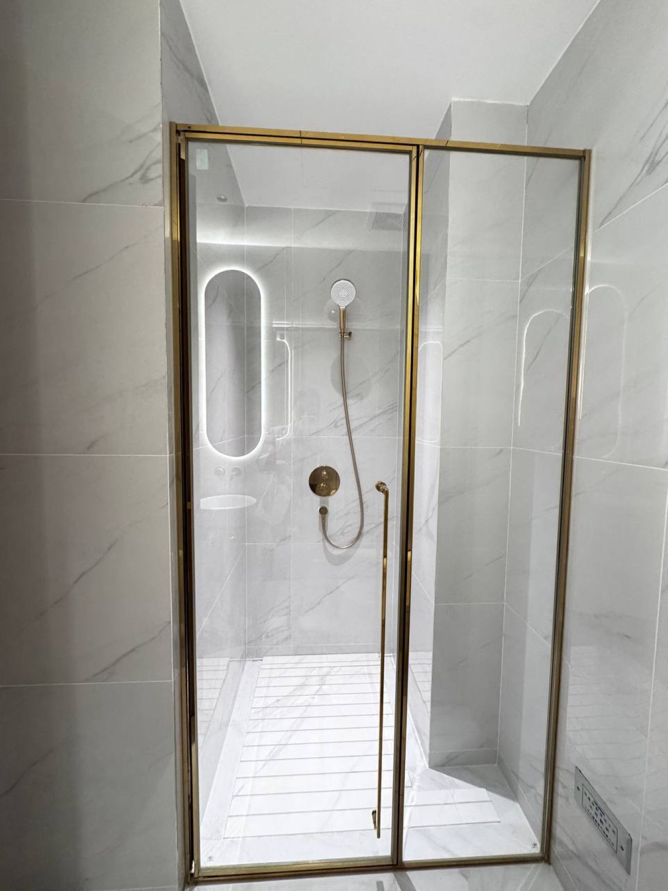 Une douche fermée par des parois de verre, avec un cadre doré, des carreaux de marbre blanc et une prise électrique visible.