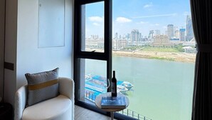 Ein modernes Hotelzimmer mit einem großen Fenster und Blick auf die Stadt, einem runden Tisch mit einer Flasche sowie einem weißen Sessel.