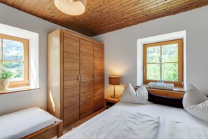 A bedroom with a wooden wardrobe, a bed with white bedding, a bedside table with a lamp, and a window with a view of greenery. - Ferienhaus Auwiese - Family Retreat (Lend)