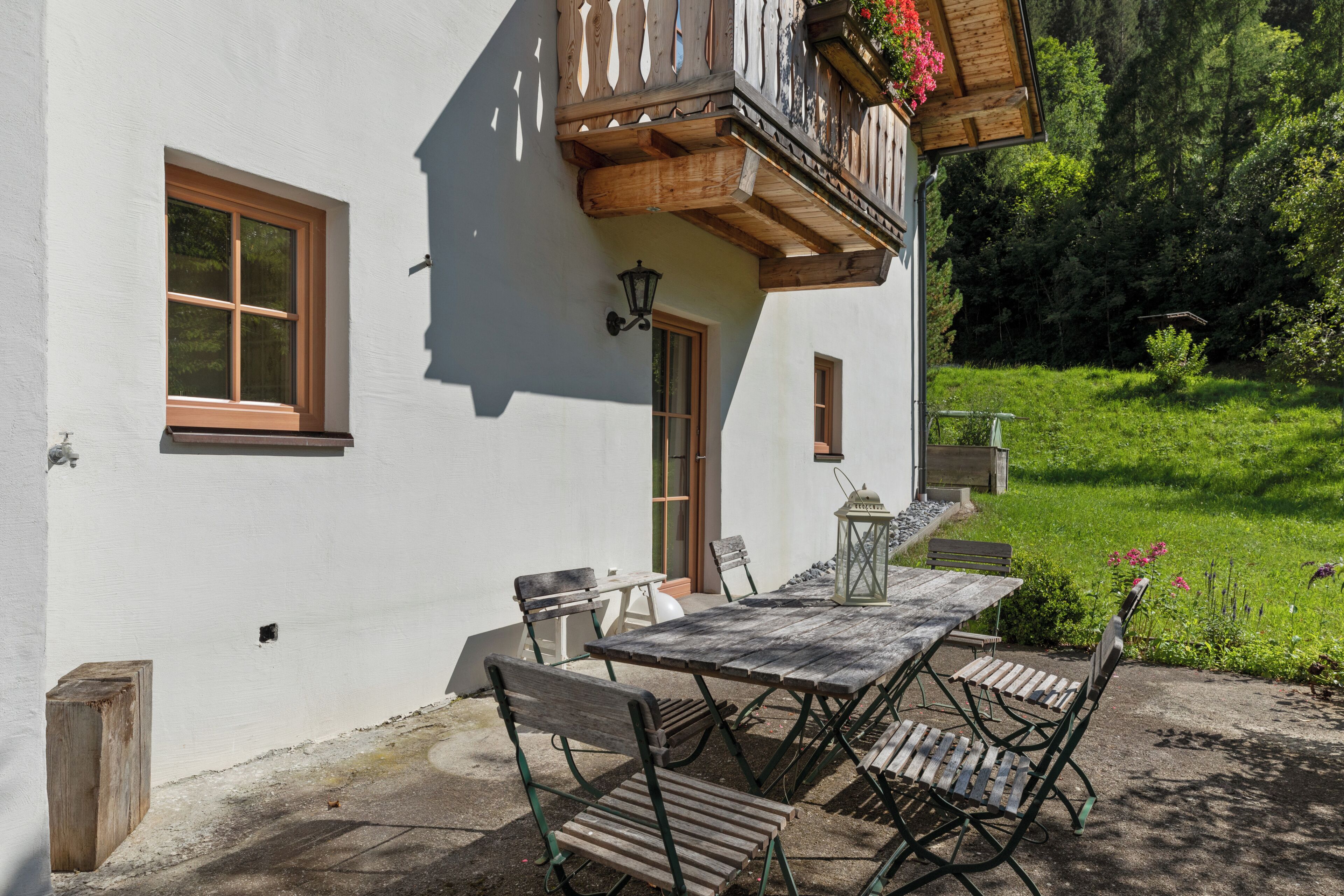 A white building with a balcony, a table with chairs, and a garden in the background.