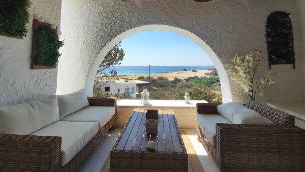Una habitación con pared blanca texturizada, techo de madera, un amplio ventanal arqueado, mobiliario de mimbre y vista a la playa.