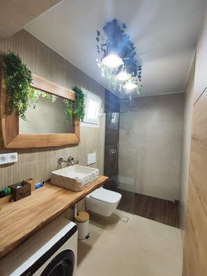 A modern bathroom with a wooden vanity, a large mirror, a built-in sink, a shower, and a hanging plant.