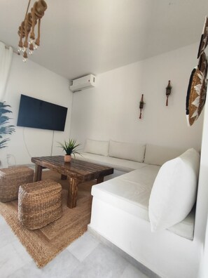 Un salon moderne doté d’un téléviseur à écran plat, d’une table basse en bois, de poufs en osier, d’un canapé blanc et d’un ventilateur de plafond.