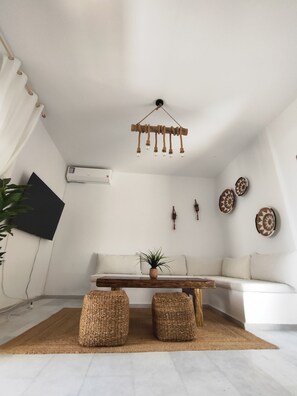 A modern living room with a white sofa, a wooden coffee table, woven poufs, a hanging light fixture, wall-mounted decor, and an air conditioning unit.