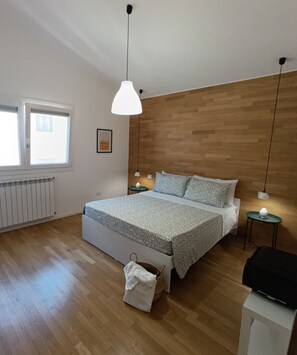 Ein Schlafzimmer mit hölzernem Kopfteil, einem Bett mit gemustertem Bettbezug, einem Nachttisch mit Lampe, einem Heizkörper und einem Fenster mit Jalousien.